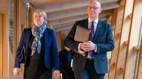 Shona Robison wearing a dark suit looking slightly off camera walking next to John Swinney, also wearing a dark suit, at the Scottish Parliament.