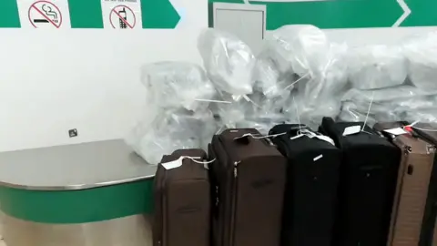 A set of brown luggage next to a silver worksurface with piles of plastic bags on it