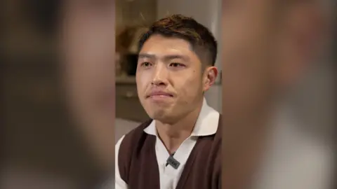 A young man looks into the distance. He is wearing a white polo shirt with a brown vest over it.