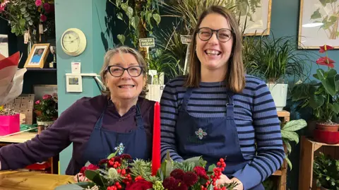 ELLEN KNIGHT/BBC Two women stood side by side and smiling. Catherine, on the left, is wearing a dark purple long sleeve top and a navy apron. Bea, on the left, is wearing a blue and navy striped top with a navy apron. They're stood behind a counter in the shop, which has teal blue walls and wooden shelves with pot plants like peace lilies on behind them. In the foreground is a Christmas candle arrangement - a tall red candle, with its base covered in green and red foliage. 