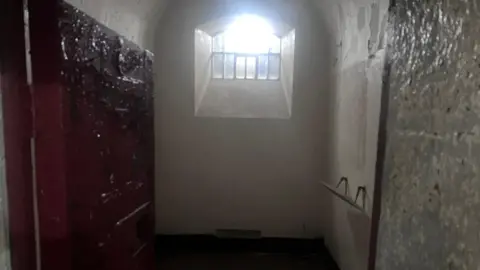 A view into a cell in Armagh Gaol. A heavy metallic door opens into a very narrow, plain room with a arched ceiling. Bright sunlight is shining through a small arched window, secured by metal bars. Paint is pealing off the walls and the door. 
