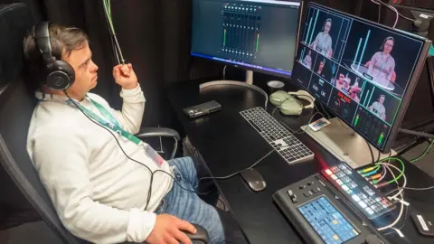 A young man sits watching screens in a television control room. He looks relaxed as he leans back but focused on what he is watching. He is dressed in a light, long-sleeved t-shirt and denim jeans, and wearing headphones and an identity card on a green lanyard. 