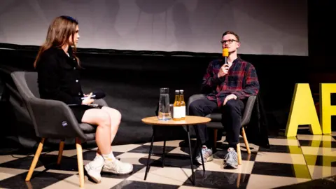 Éanna Mac Cana A wide shot of Éanna Mac Cana taking part in an on stage interview. A woman with long dark hair and wearing a dark denim jacket is holding a microphone as she sits in a grey chair. There is a small circular tablet holding two glass bottles and a carafe of water. Éanna is wearing small, dark framed glasses and is holding a microphone with a orange head on it to his mouth. He is wearing a navy and red checked flannel shirt, a pair of dark jeans and grey trainers. The stage is a black and white checked pattern and they're sitting in front of a large projector screen.