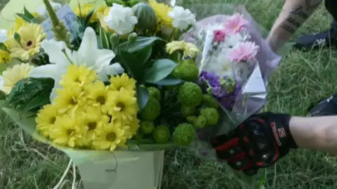 A bunch of yellow and white flowers in a bouquet with a pair of gloved hands adding some pink and purple flowers