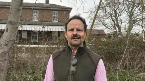 James Mallinder James Mallinder wearing a dark vest over a pink shirt standing in front of some bushes with Melton railway station in the background.