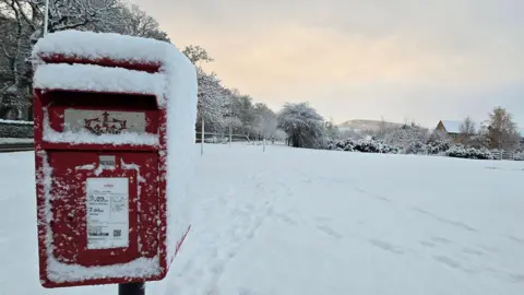 BBCWeatherWatchers/Emma A postbox with snow on its top and sides in the foreground and snow-covered land in the background. 