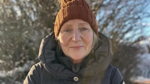 Sarah Johnson in a brown woollen hat and a large puffa-style jacket, standing in front of a tree covered in snow