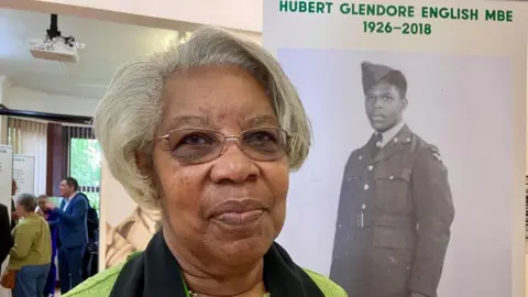 An older woman with short grey hair and darkly tinted glasses stands in front of a sign with a black and white image of a soldier. Above the full-length photograph is text reading Hubert Glendore English MBE 1926 - 2018. 