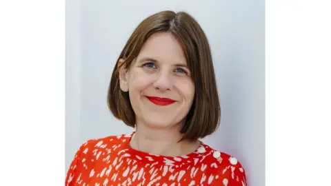 Beth Moseley Rebecca Atkinson wearing a red and white blouse. She has short brunette hair and is wearing red lipstick. She is looking directly at the camera and smiling.