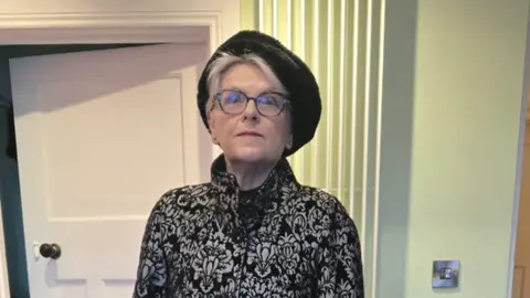 FELICITY HOUSTON A woman with fair hair is standing wearing a black and grey patterned coat and a black hat. 