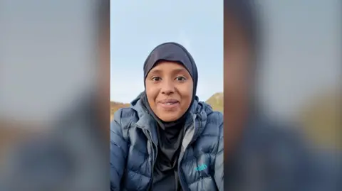 Amina Hassan smiles at the camera. She is wearing a blue rain coat, and a black hijab. There are hills behind her and a blue sky.