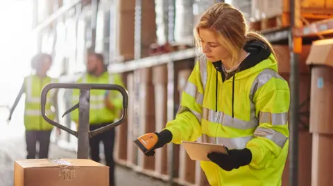Young woman with long blonde hair tied back in a loose ponytail wearing a high vis jacket in a warehouse scanning a parcel on a trolley. There are shelves of parcels behind her and two colleagues also wearing high vis walking towards her.