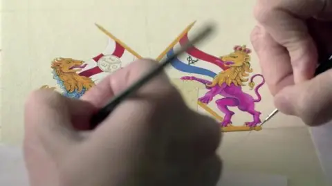Alex Dunlop/BBC An intricate painting of small gold, pink and blue lion statues, holding flags - with two hands painting them.