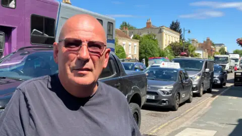 A man with sunglasses standing infront of a high street packed with cars