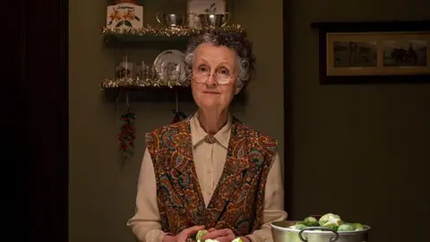 BBC/Neal Street Productions Miss Higgins, played by Georgie Glen, peeling sprouts in the kitchen in a white blouse and apron