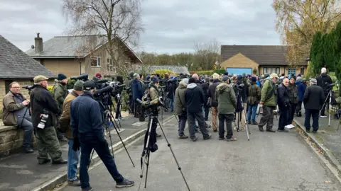 BBC/Tom Ingall Small residential street with a large crowd of mostly men holding tripods and binoculars dressed in warm coats looking towards one spot