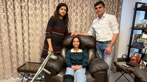 A man and a woman stand either side of a brown leather armchair their 12-year-old daughter is sat on in their living room. Her crutches are resting on the arm of the armchair. She is smiling but her parents have quite a serious expression on their faces.