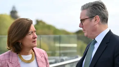 Eluned Morgan, who has bobbed brown hair and is wearing a white t shirt, pink check blazer and gold necklace is looking at Sir Keir Starmer, who has short grey hair and is wearing a dark suit, white short and green-blue tie.