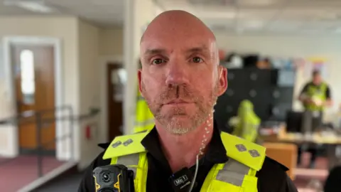 West Midlands Police A police officer with a small white beard wearing a high visibility jacket and body camera in an office