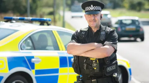 Kevin O'Mara A man in a black police uniform and black and white cap is stood alongside a liveried police car. His arms are folded and he is smiling