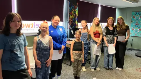 Jon Wright/BBC Eight young women and girls stand in a line inside a music room. Some are holding sheets of paper.