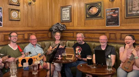 A group of men and women are sitting on a green leather sofa holding ukuleles. They are laughing and sitting next to wooden circular tables, with pints of beer and lemonade. 