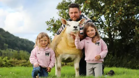 Michael Davies kneels behind a sheep. He is wearing a black and red and white stripey jumper and has short brown hair and stubble.One of his young daughters, who looks about five, kneels next to the sheep in a pink jumper and blue jeans. She has curly blonde hair. On the right of the sheep Davies' older daughter, who looks to be about seven or eight wears a matching pink hoodie and grey jeans. She has long brown hair. She is holding the sheep's head. Bind them in the background a tree and bushes are visible. 