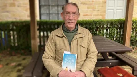 A picture of Tom holding his book, The Penguin Lessons. He is wearing a cream coloured coat and holding a blue book. He is wearing glasses.