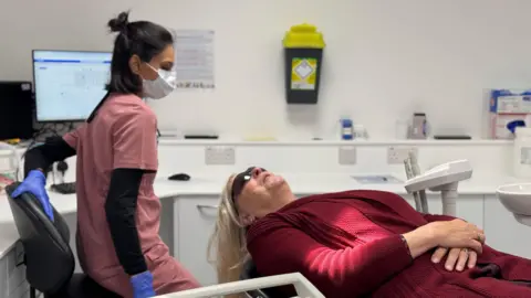 BBC Heather, wearing a red sweater dress, is laid on a dentist chair, wearing black glasses with her arms crossed across her abdomen. A woman in pinkk scrubs and a white mask looks down at her. In the background there is a computer and a workbench.