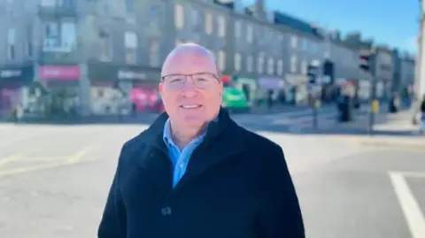 Bob Keiller stood on Union Street on a sunny day. He is wearing a blue shirt, black jacket and has glasses.