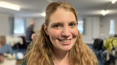 Vikki has long light brown hair which is tied back but also flows over her shoulders. She has brown eyes. She is smiling for the camera.