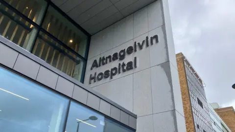 A close up of a sign outside Altnagelvin Hospital, which says the name of the building in large lettering. It is a tall building with a mix of facades including brown brick and grey cladding. 