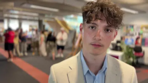 BBC/NATHAN TURVEY 18-year-old James Pearson, who has brown curly hair and is wearing a cream jacket and blue shirt, with people standing behind him.
