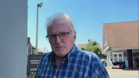 John is smiling at the camera and he looks a little cheeky. He is standing near a CI Coop store and there is a road, street light and CI Coop parking sign behind him. He is wearing a pair of glasses and a blue checked shirt and he has short white hair.