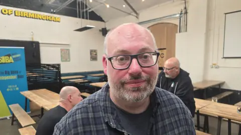 A man with white hair and a beard, wearing black glasses and a checked shirt, smiles at the camera. He stands in front of several benches inside a room.