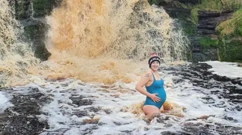 Kellie Tarr Kellie Tarr is wearing a blue swimming costume and is pregnant, she is standing in front of a waterfall