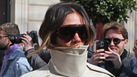 Getty Images Victoria Beckham in a beige funnel-neck jacket and sunglasses leaving her hotel during Paris Fashion Week