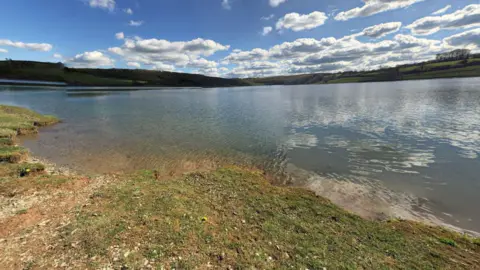 Wimbleball Lake images taken from Google Maps showing the water with hills in the distance, and a gravel beach in the foreground