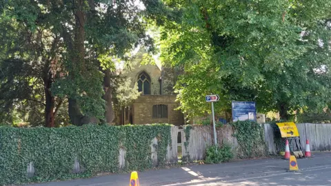 Outside St Thomas the Martyr church on a sunny day. There is a yellow road sign redirecting traffic in front of its yard. Some yellow cones can be seen on the road as well.