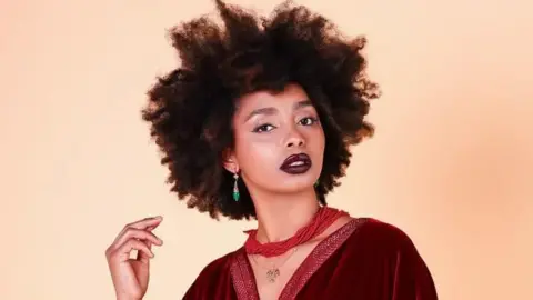 Houda Fonone / BOCHIC Head and shoulders shot of model and content creator Houda Fonone, wearing a dark red top, a necklace of strands of red beads, and green earrings.