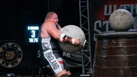 Ben Musgrove Dean Mason - a man with broad shoulders and muscles - is wearing a grey shirt that says England with a lion down the side of it. His face shows he is straining while lifting a large circular grey ball which looks like concrete. A brown barrel next to him has another large grey ball on top of it.
