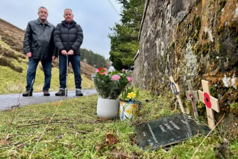 The plaque reads: Maj D Cunningham, S Sgt Hosker T, Pte Thomson J - who were killed whilst carrying out their duties on 17 Jan 1985, Lest we forget.
There are three small wooden crosses behind the plague. Mr Anderson and Mr Allan are in the background of the picture and the plaque is in the foreground.