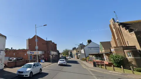 Google Street view of Oundle Road with a church building on the right and the plot of land on the left and cars on the street