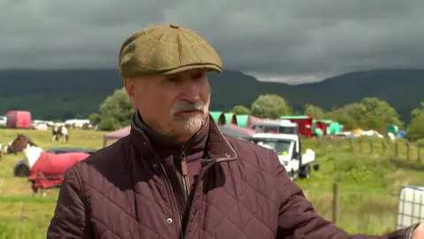 Billy Welch has a white moustache . He is wearing a burgundy top and jacket and a green tweed baker boy hat.