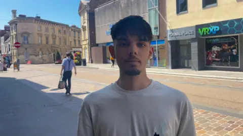 The image shows a young man in the centre of the picture standing on the pavement of Gloucester highstreet, shops can be seen in the background and there is a man carrying a bag in the back of the picture.