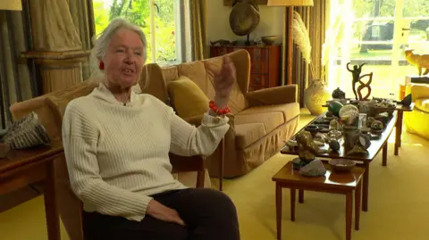 Mary Moore, daughter of Henry Moore, sat on a light coloured sofa wearing a ream jumper and withher white hair in a ponytail