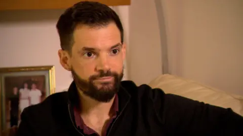 Family handout Alexander Robinson, with short dark hair and a beard is wearing a dark brown jumper, sitting on a sofa looking to camera with a neutral expression, with a family photo blurred in the background.
