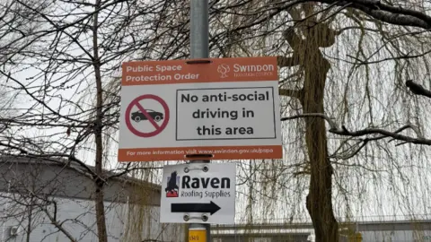 Swindon Borough Council A sign reads 'no anti-social driving in this area' with a car inside of a no entry symbol. It has Public Space Protection Order and Swindon Borough Council's logo on it. Bare trees and an industrial building are visible behind the sign.