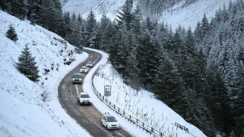 An overhead shot looking down at the Snake pass road in the Peak District. Cars travel along the road surrounded by snowy trees. 