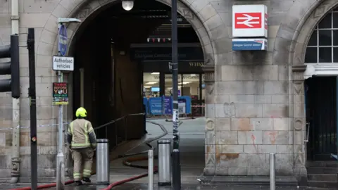 PA Media Firefighter in protective gear standing outside an arched entrance to Glasgow Central Station, with hoses on the wet pavement and cordon tape blocking access. A rail station sign is mounted on the stone wall above the arch.